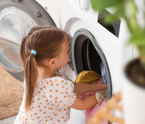 Little,Girl,Putting,Laundry,In,Washing,Machine,At,Home