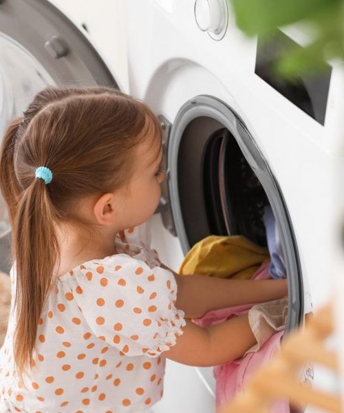 Little,Girl,Putting,Laundry,In,Washing,Machine,At,Home